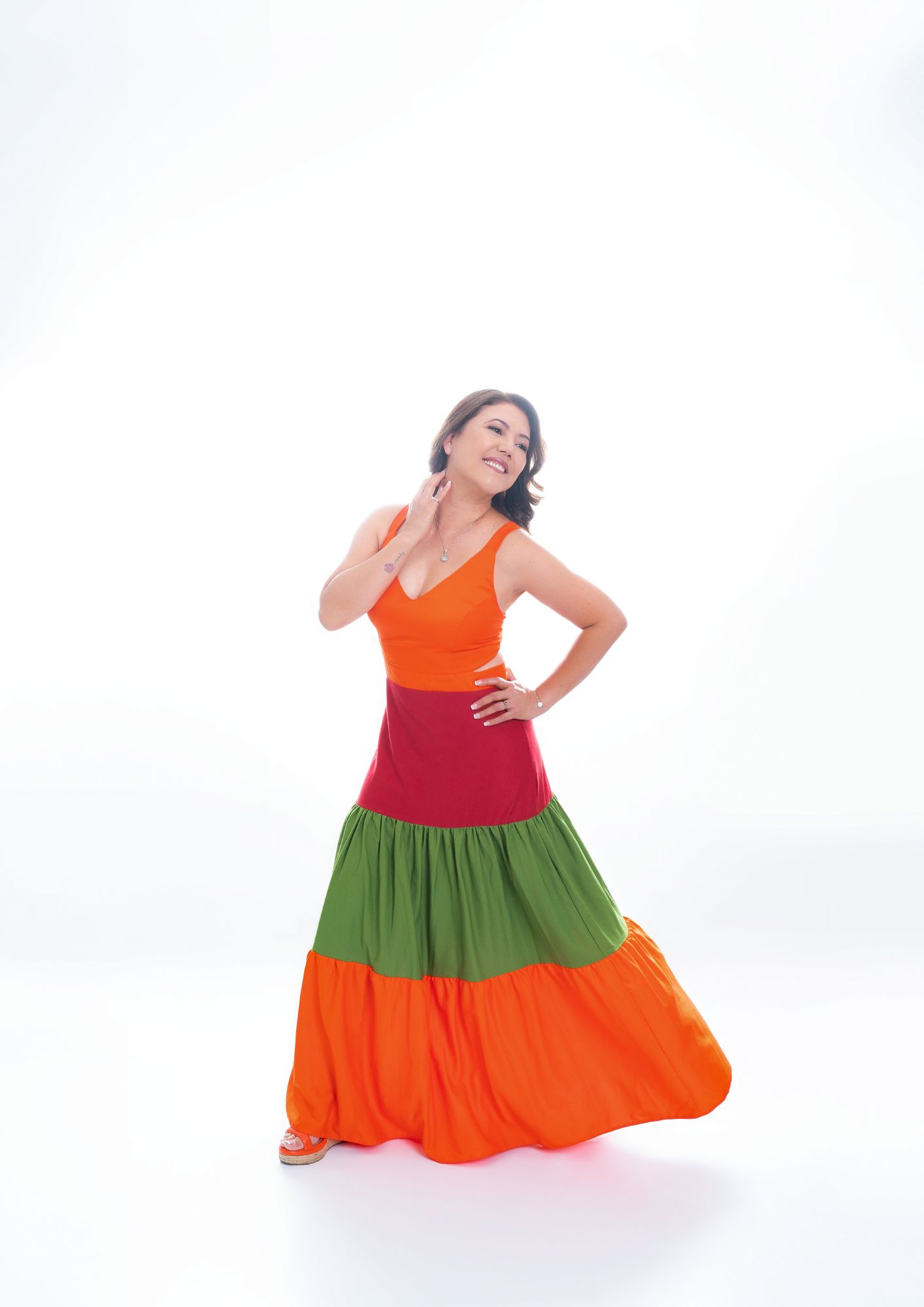 Woman in a bright orange and green tiered maxi dress dancing against a white studio backdrop, smiling with one hand on her hip and the other near her neck.