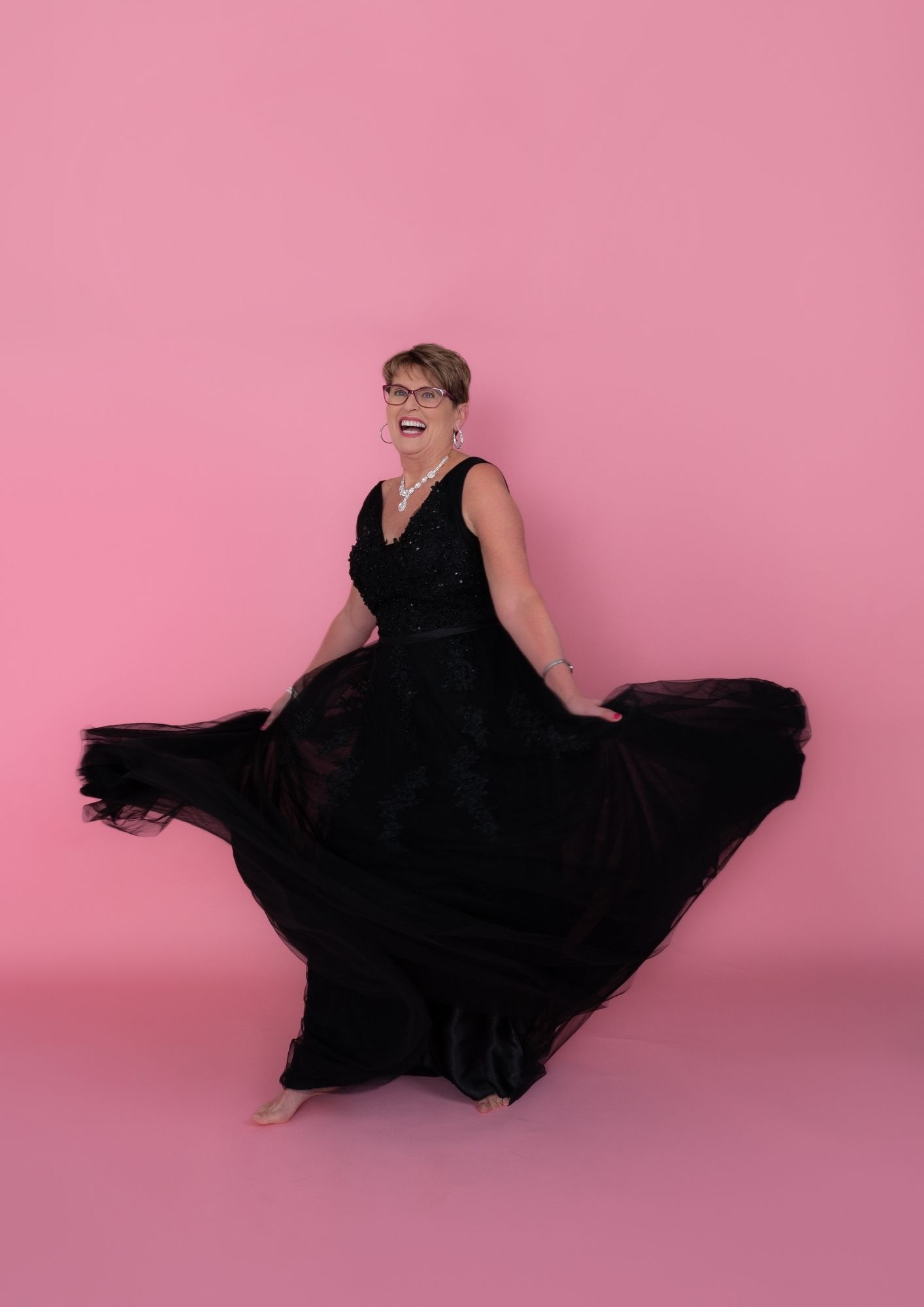Older woman in a sequined black gown spins, pink backdrop behind her, smiling widely.”] ,