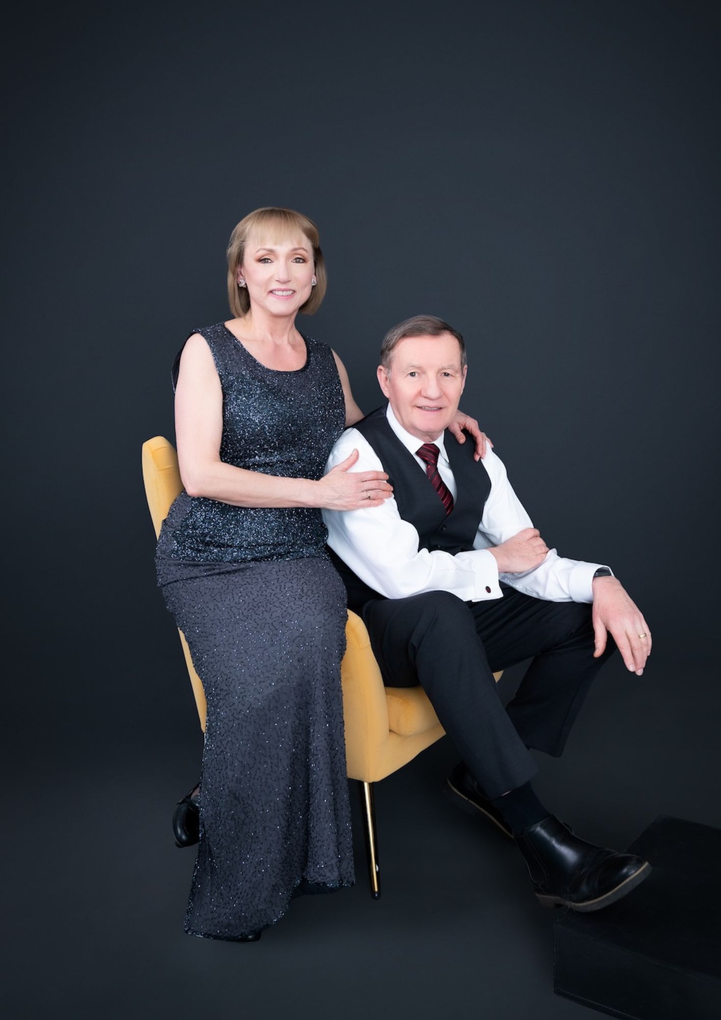 Older couple in formal attire posing together against a dark backdrop; woman standing with hand on a seated man, both smiling.