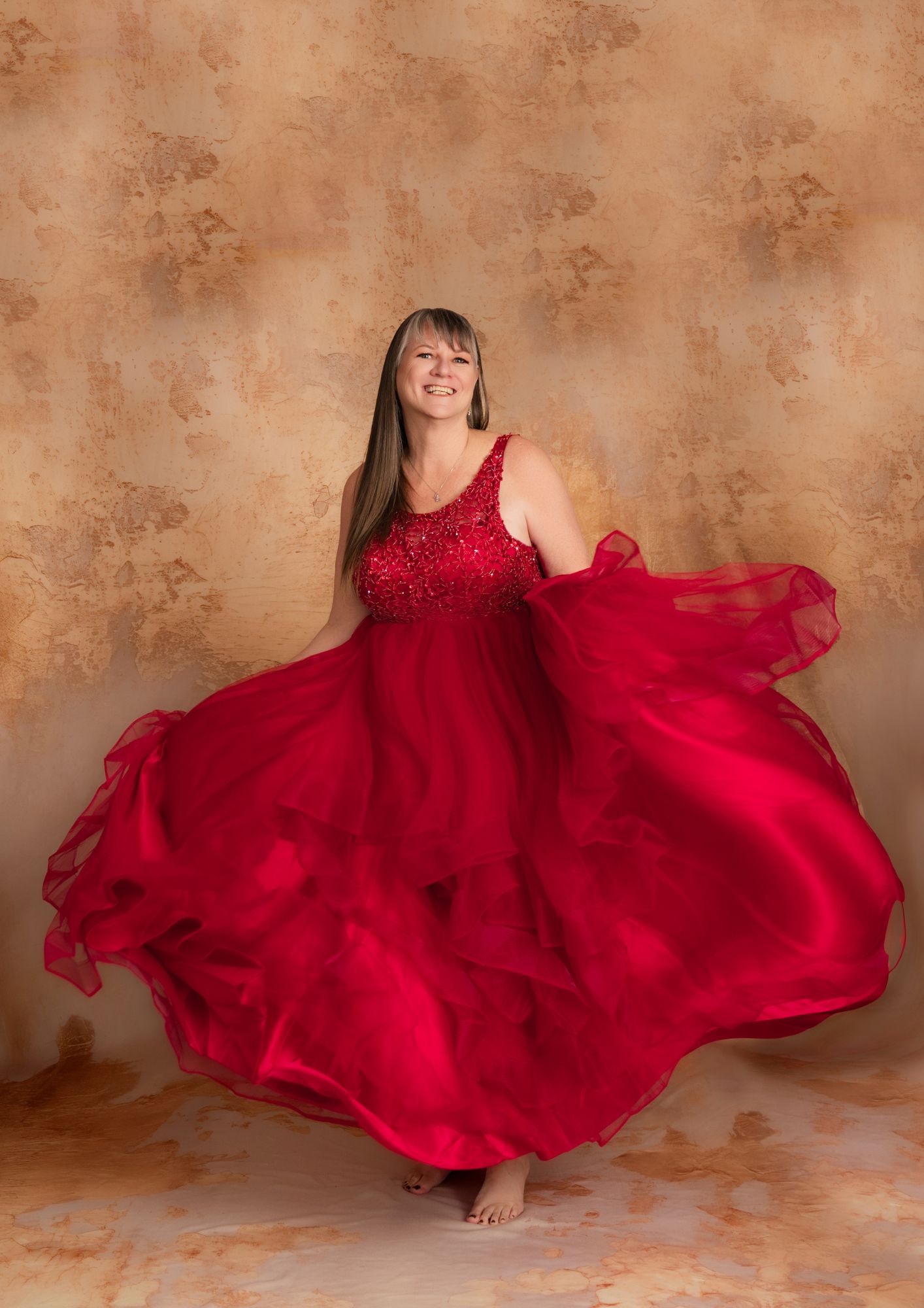 Woman in a red sequined gown twirls, smiling, with a large flowing skirt against a textured beige backdrop.